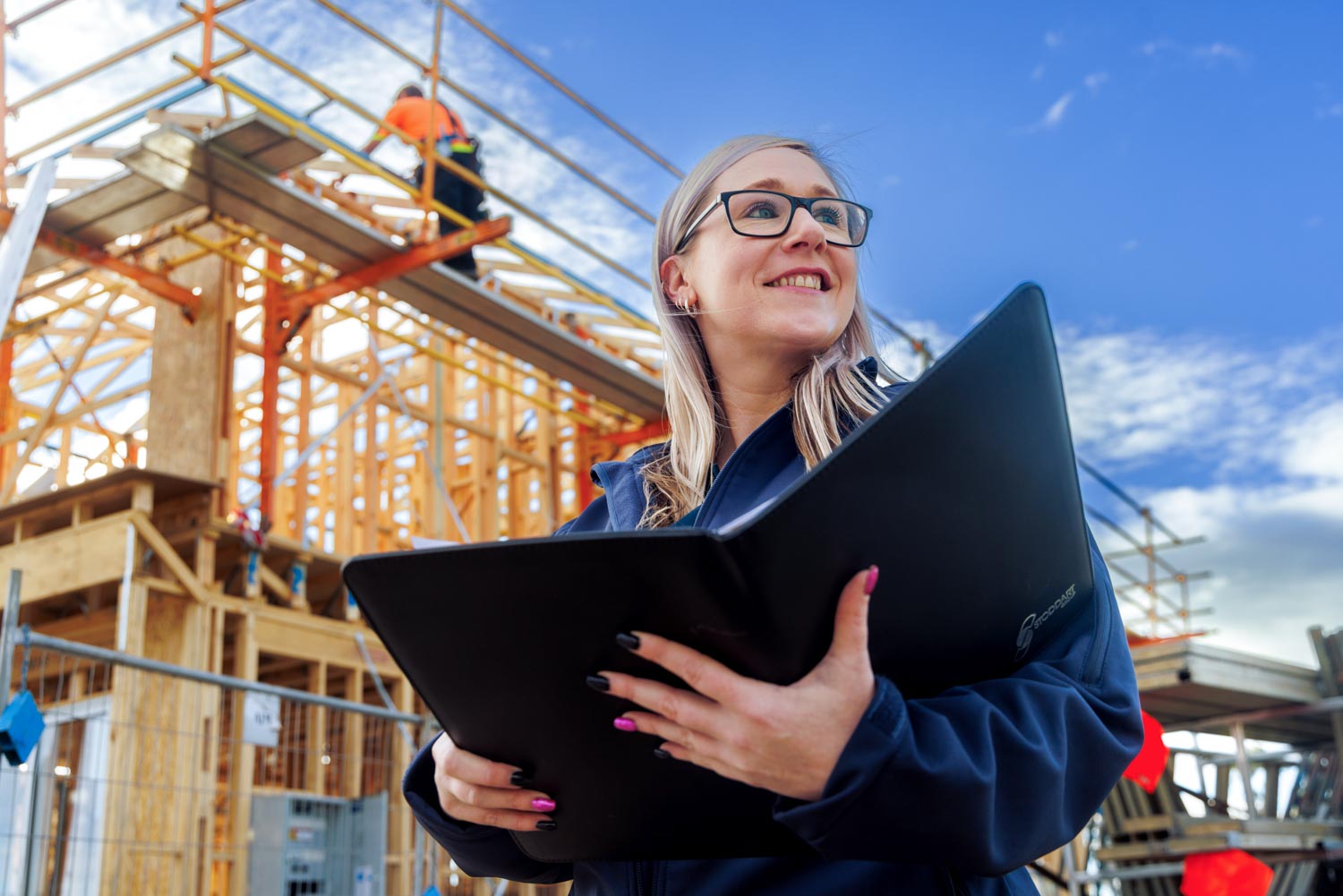 Smiling woman with glasses holding a folder in front of a wooden house under construction.