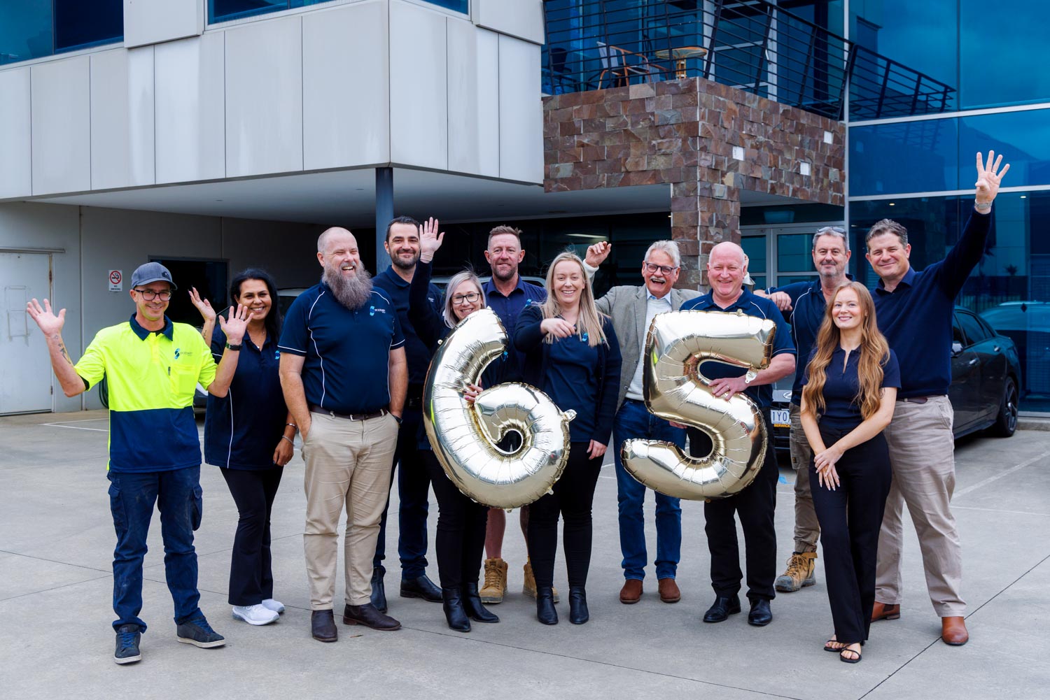 Group of twelve people standing outside a building holding gold balloons shaped as numbers 6 and 5, smiling.