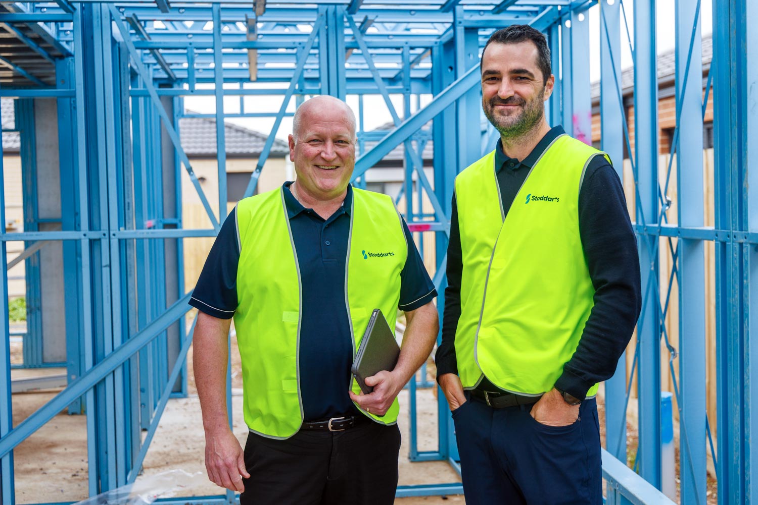 Two men in yellow safety vests standing inside a blue metal frame structure at a construction site.