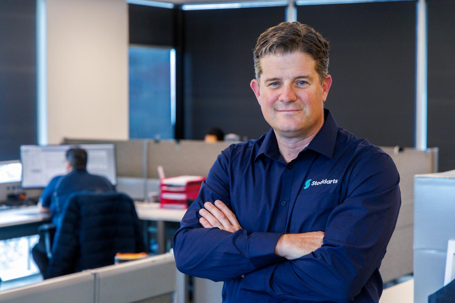 Man in blue shirt with Stoddarts logo stands with arms crossed in an office cubicle area.