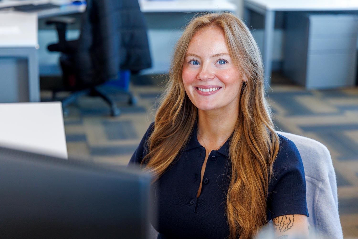 Smiling woman with long hair in navy shirt sitting in an office with desks and chairs.
