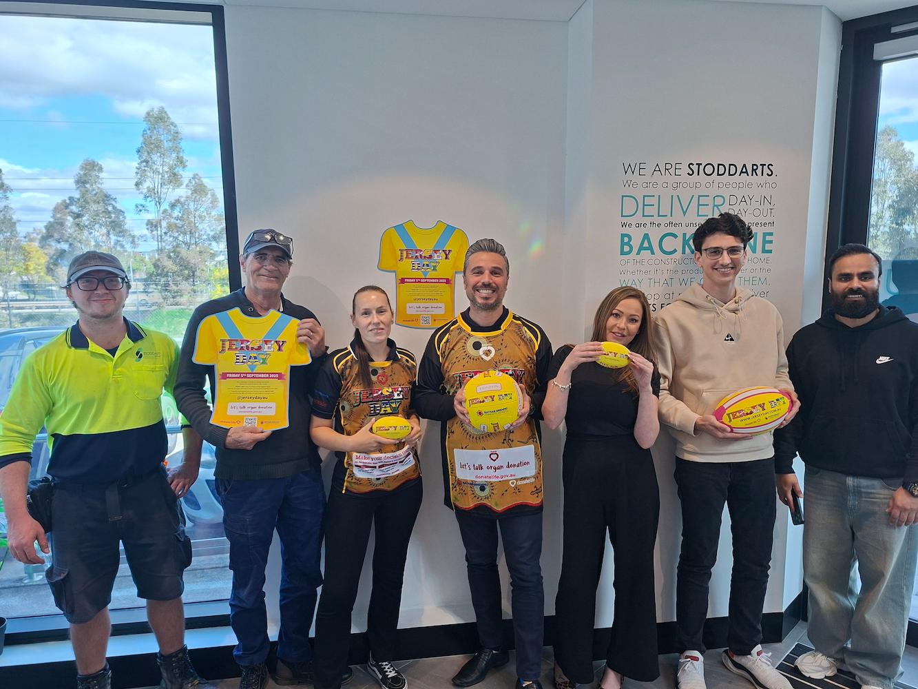Seven people indoors holding yellow Jersey Day items, standing under a sign about Stoddarts.