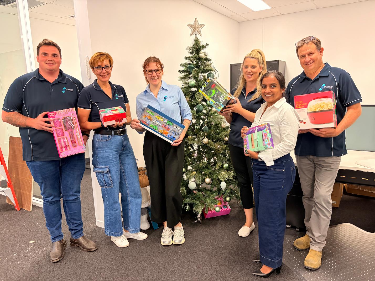 Six people in office near decorated Christmas tree holding various gift boxes and smiling.