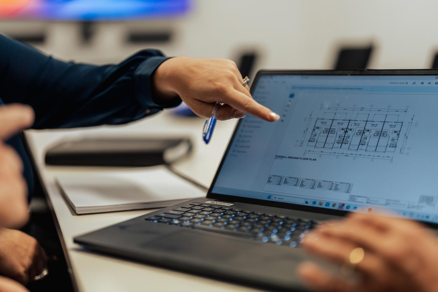 Person pointing at architectural floor plan displayed on a laptop screen in a meeting room.