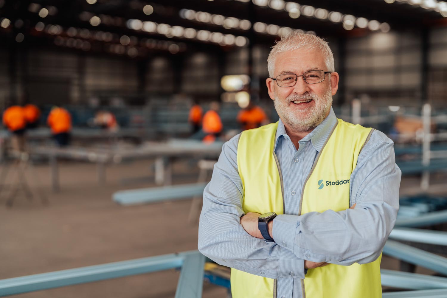 Smiling older man with glasses and beard in a high-visibility vest inside a busy industrial warehouse.