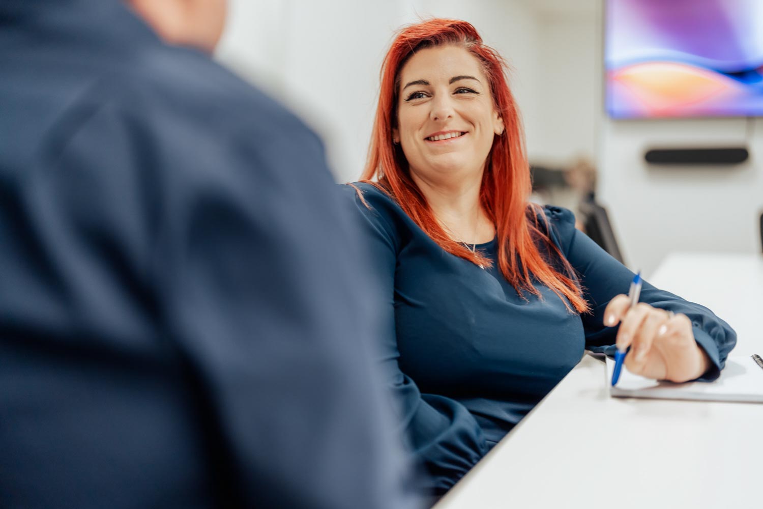 Smiling woman with red hair holding pen, sitting at a white table in conversation.