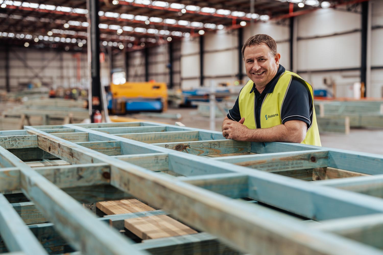 Smiling man in a yellow vest leans on wooden framework inside a large warehouse.