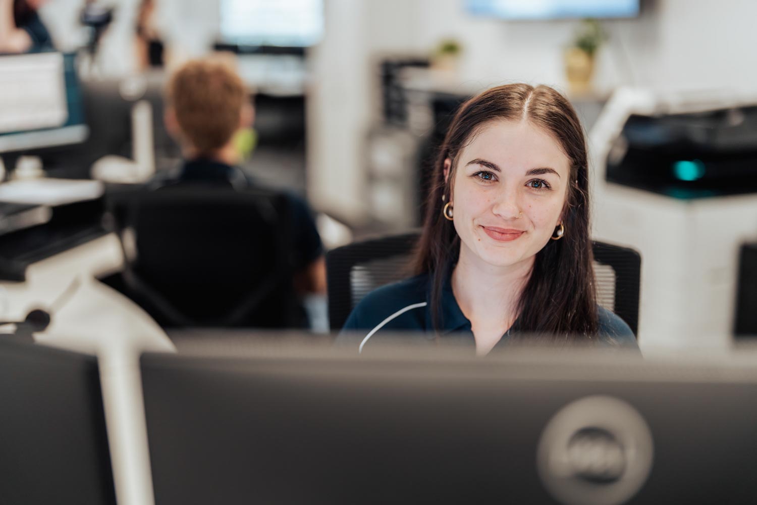 Smiling young woman with dark hair and gold earrings sitting at a computer in an office.