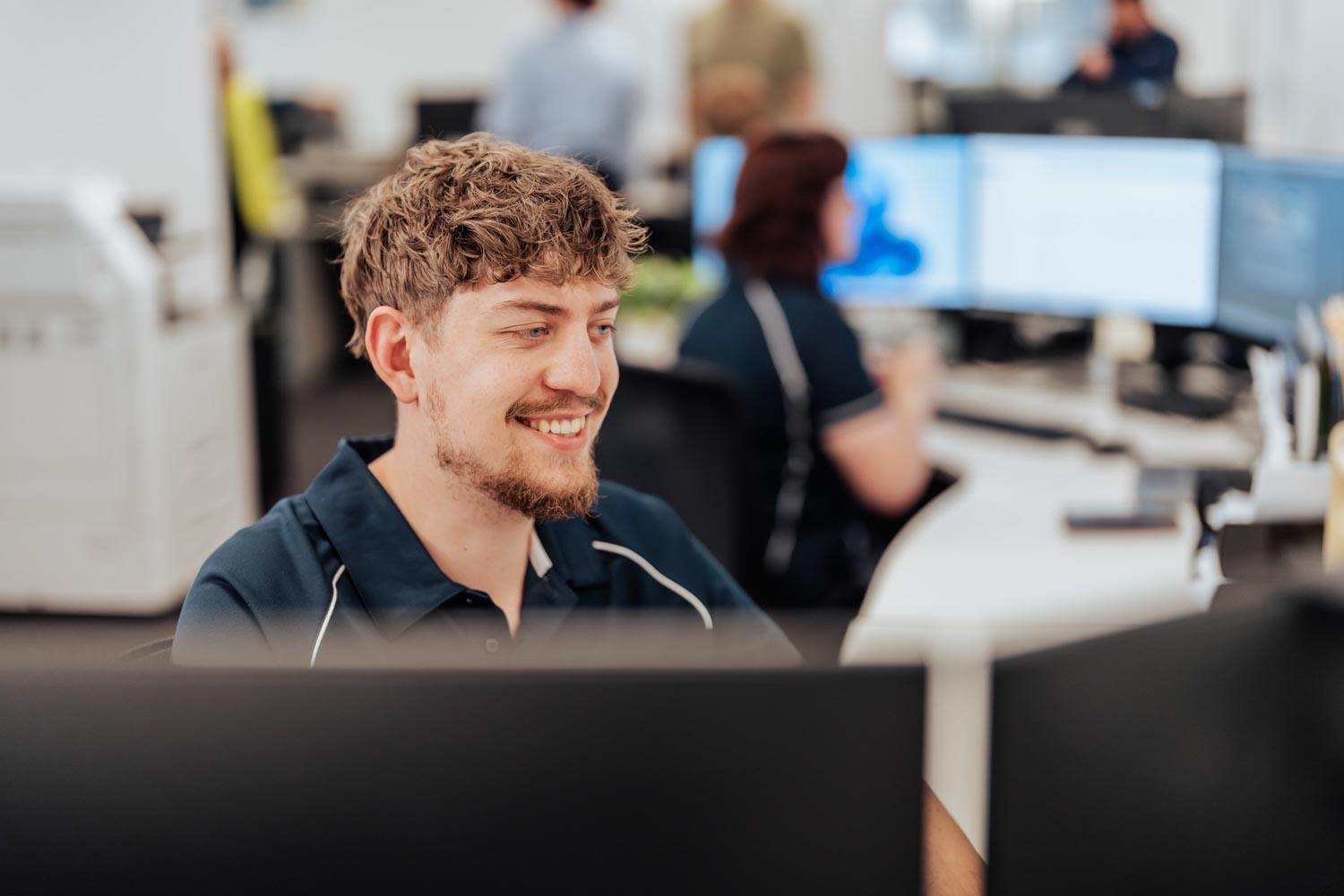 Smiling man with curly hair working at a computer in a busy modern office.