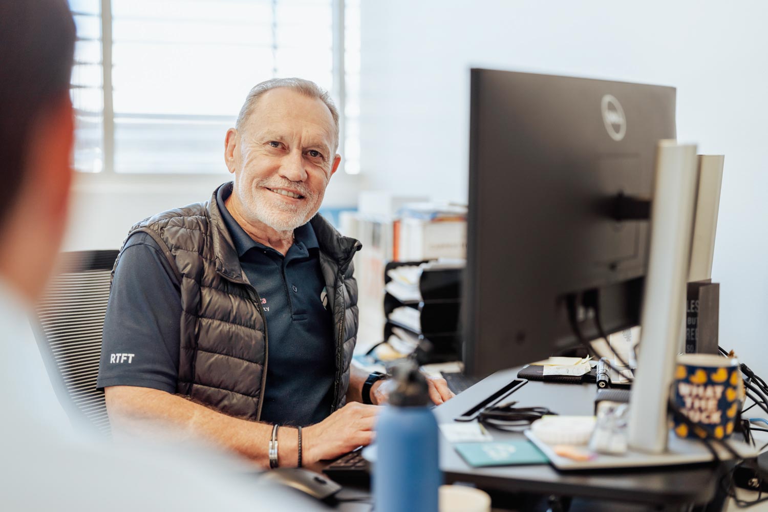 Smiling older man in black vest typing on a keyboard at a desk with a computer monitor in an office.