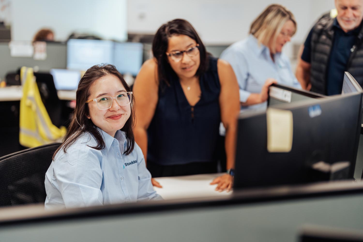 Smiling woman with glasses sitting at a desk with coworkers standing behind her in an office.