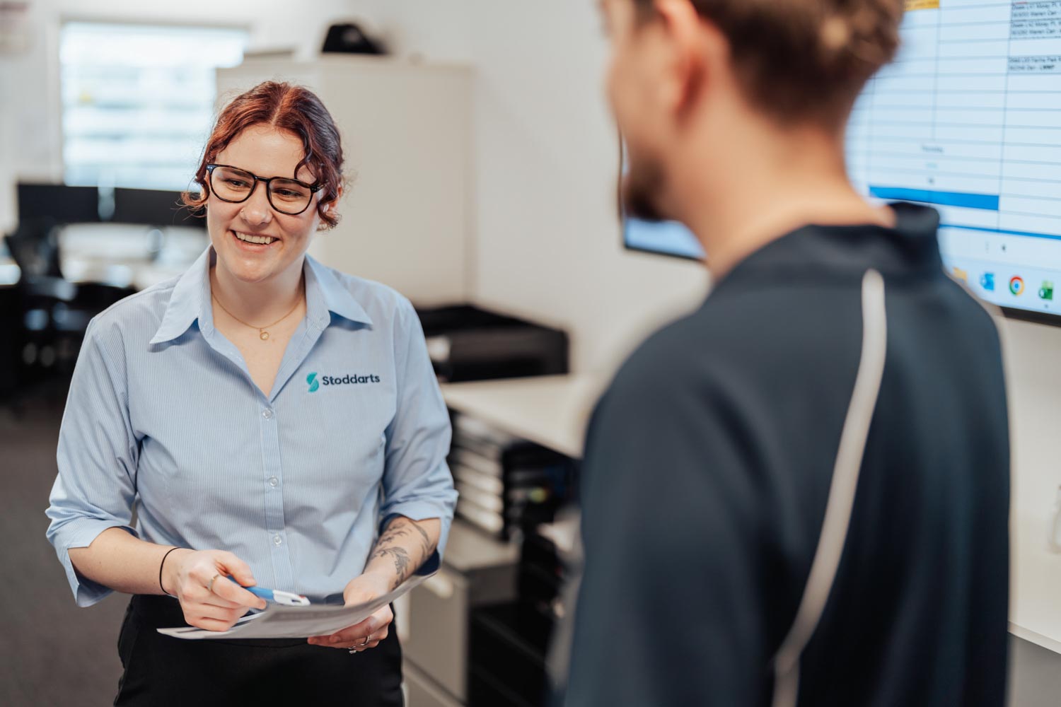Woman in Stoddarts shirt holding documents and smiling while talking to a man in an office.