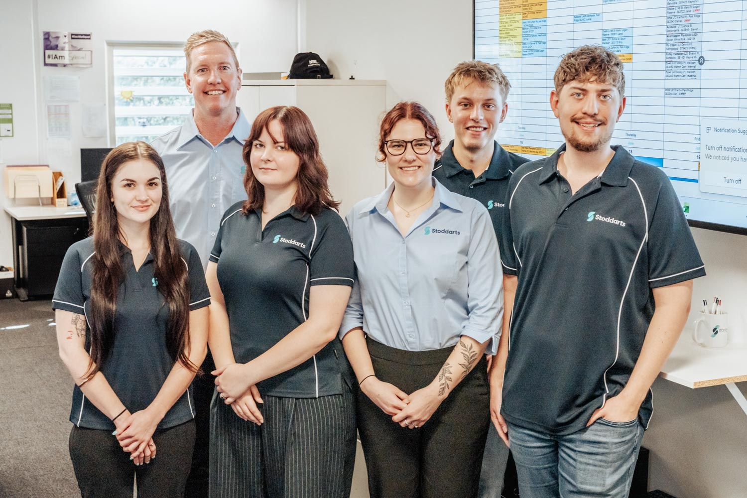 Six smiling office team members wearing Stoddarts logo shirts standing in a modern office space.