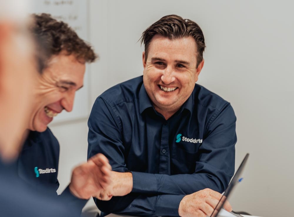 Two men in navy Stoddarts shirts laughing and chatting during a meeting in a bright office.