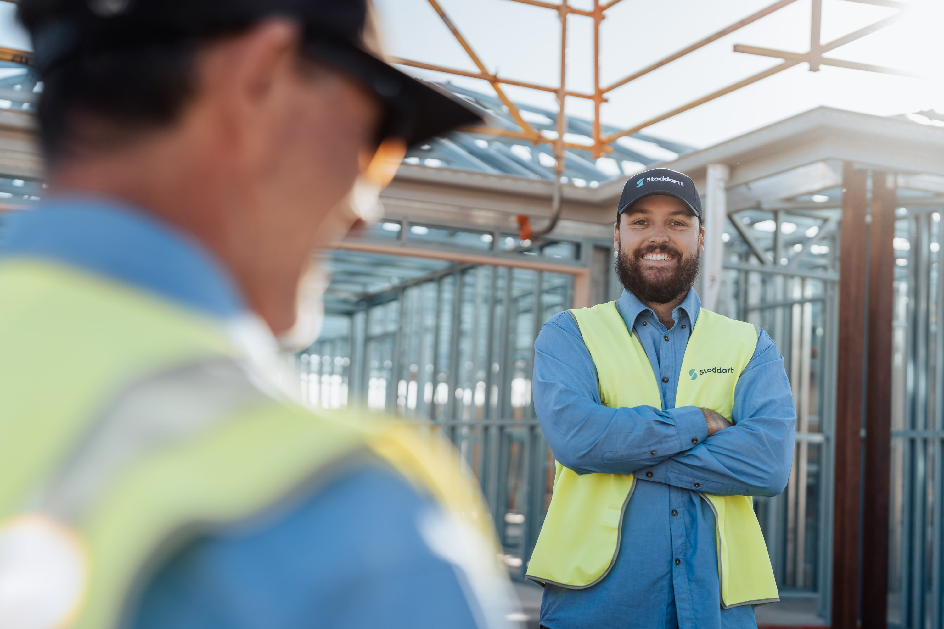 Smiling construction worker wearing a Stoddart cap and safety vest at a building site.