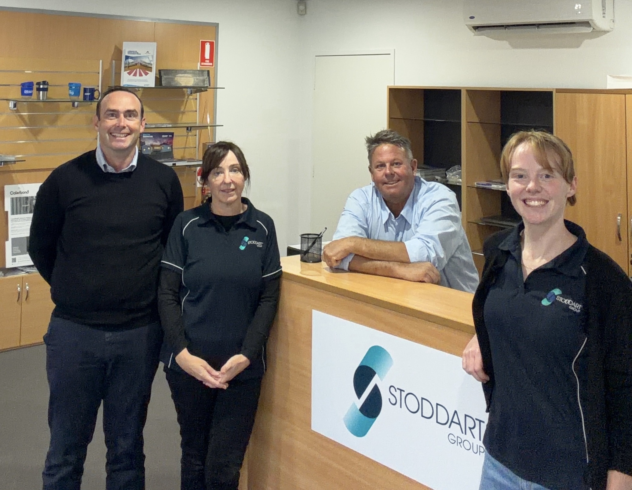 Four people smiling inside an office near a desk with a Stoddart Group sign.
