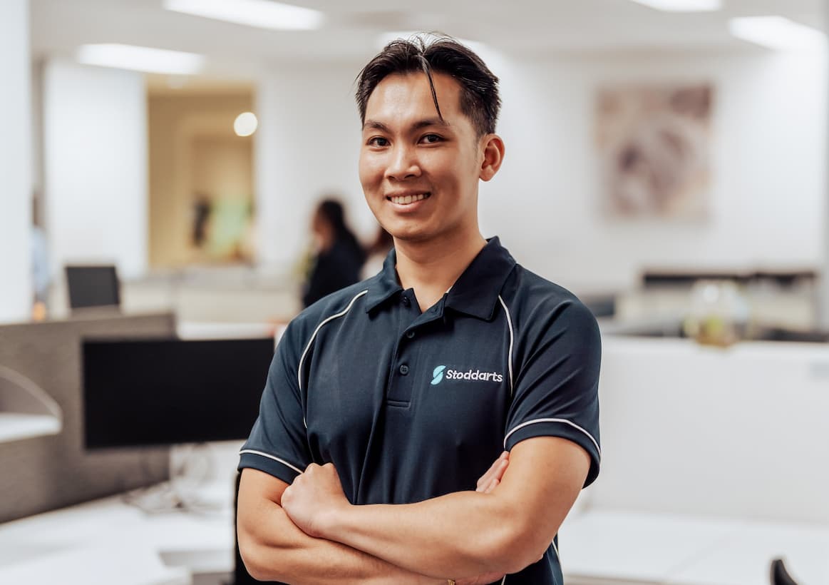 Smiling man in a navy Stoddarts polo shirt with arms crossed in a bright office space.