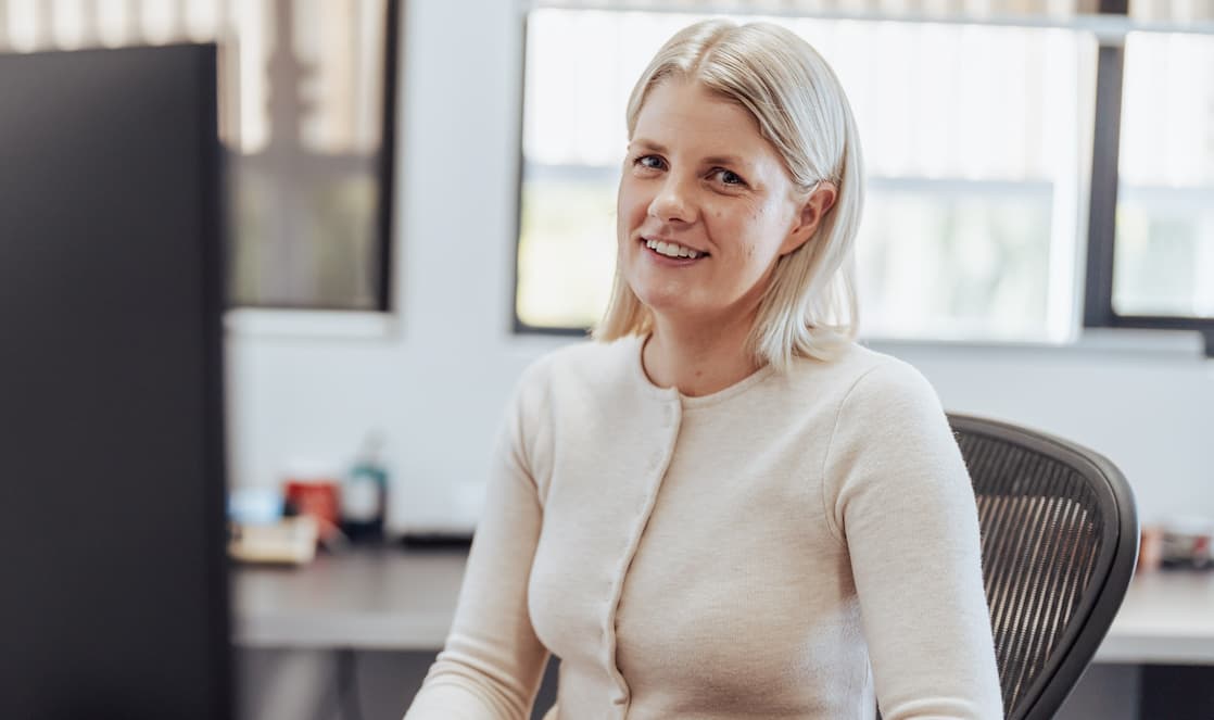 Smiling woman with blonde hair sitting on office chair near desk with computer screen.