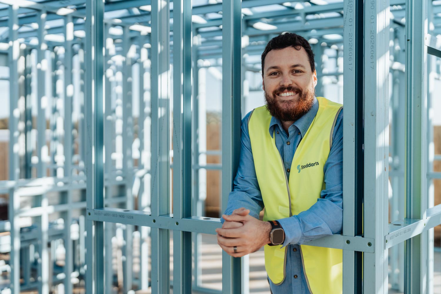 Smiling man in yellow Stoddarts vest leans on metal framing at a construction site.