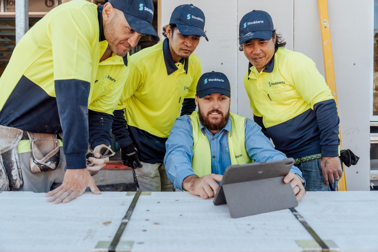 Four construction workers in Stoddarts uniforms gathered around a tablet on a worksite table.