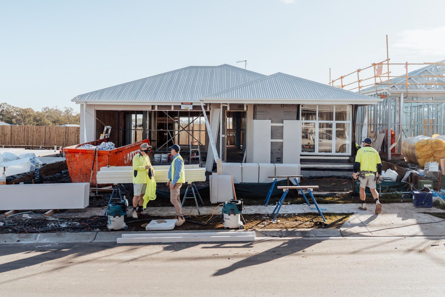 Three construction workers in yellow shirts at a partially built house with building materials outside.