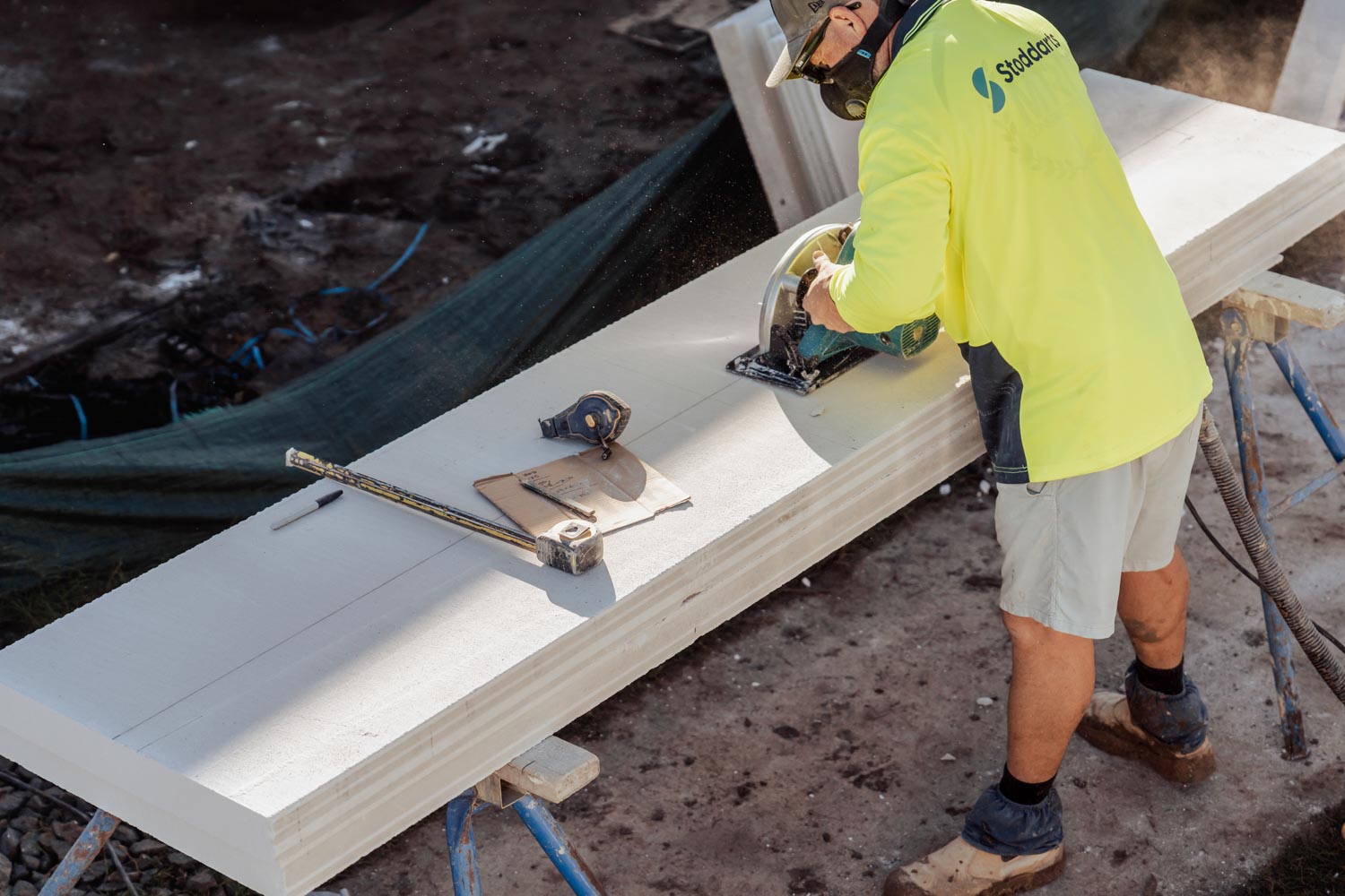 Worker in high-visibility shirt using circular saw to cut a large board on sawhorses outdoors.