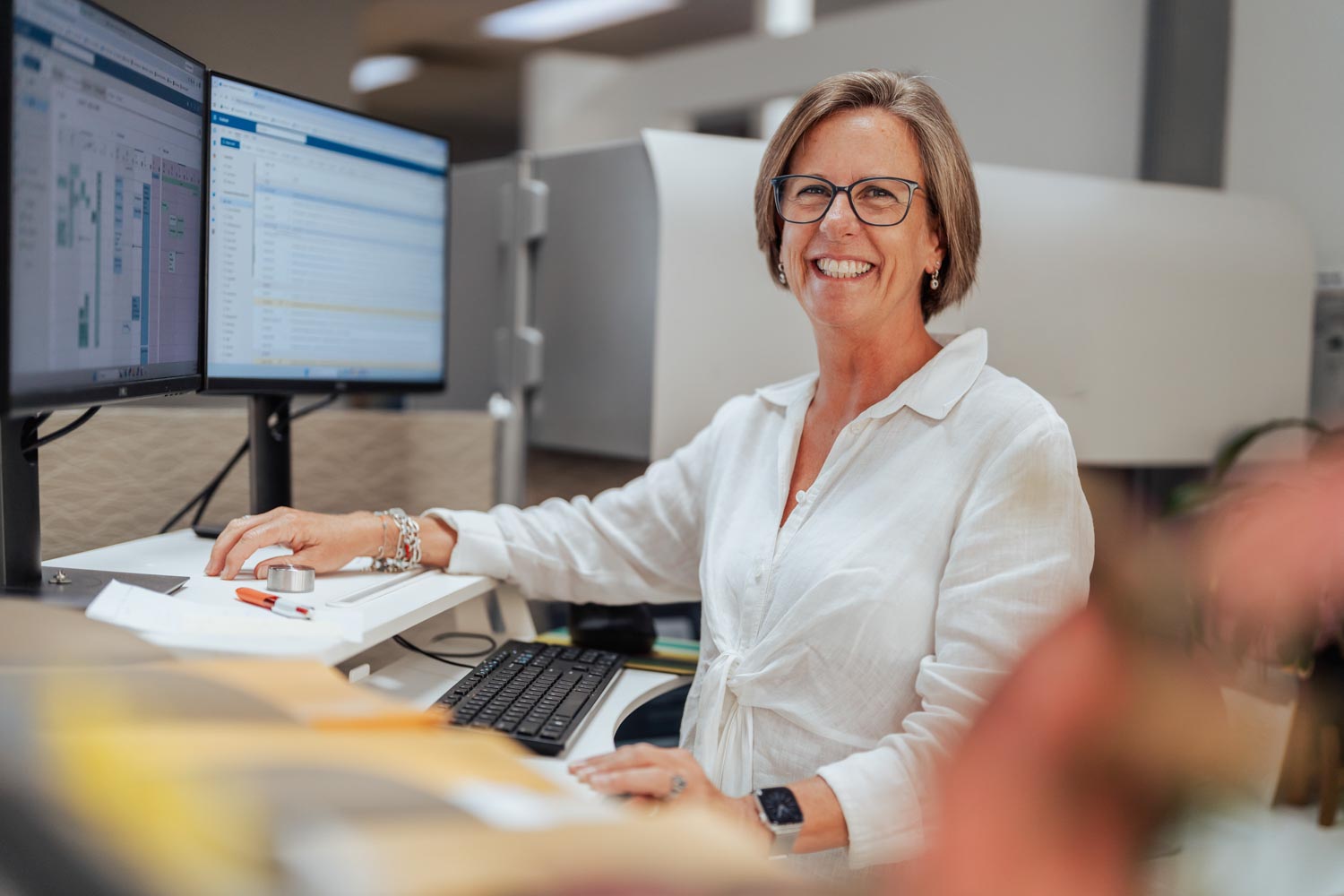Smiling woman with glasses working at a dual-monitor computer desk in an office.