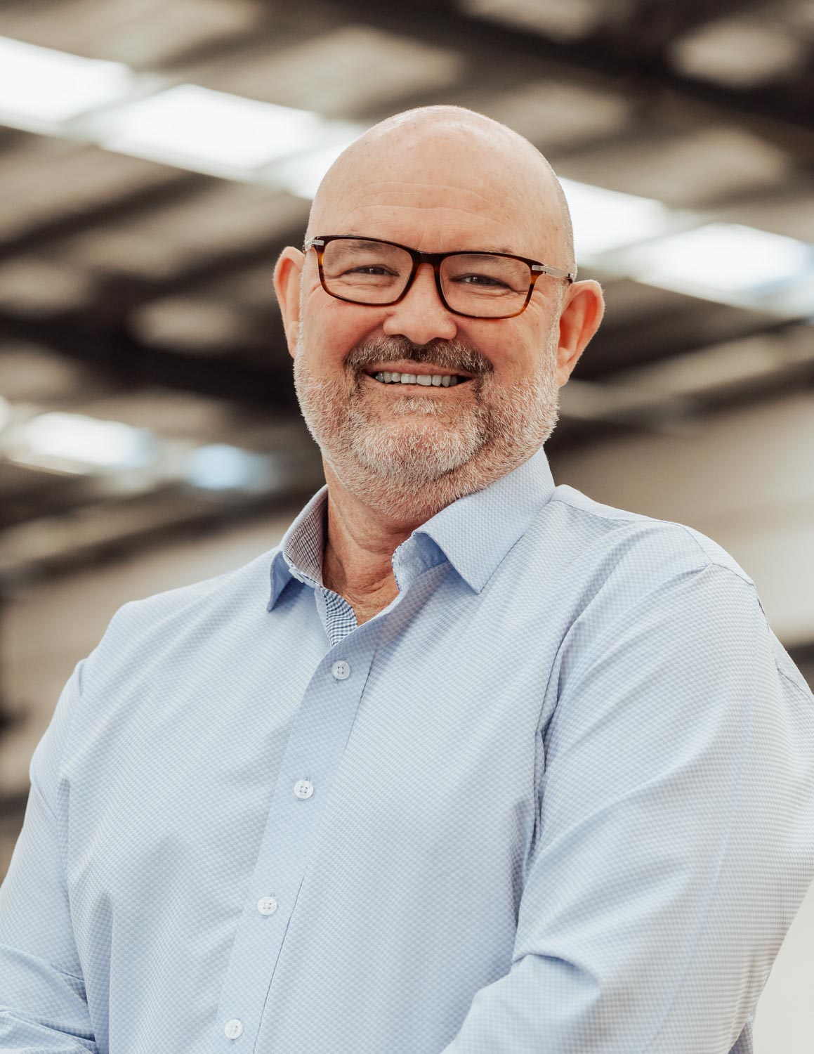 Smiling bald man with glasses and beard wearing a light blue shirt in a bright industrial setting.