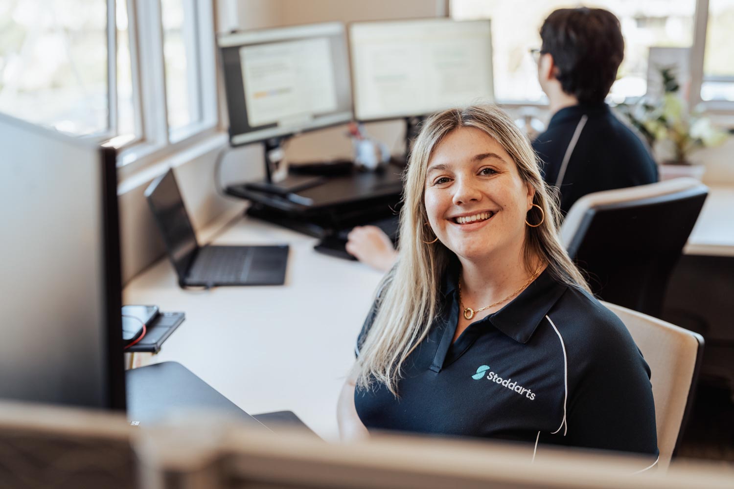 Smiling woman wearing a black Stoddarts shirt seated at a desk with computer monitors and a man working in the background.