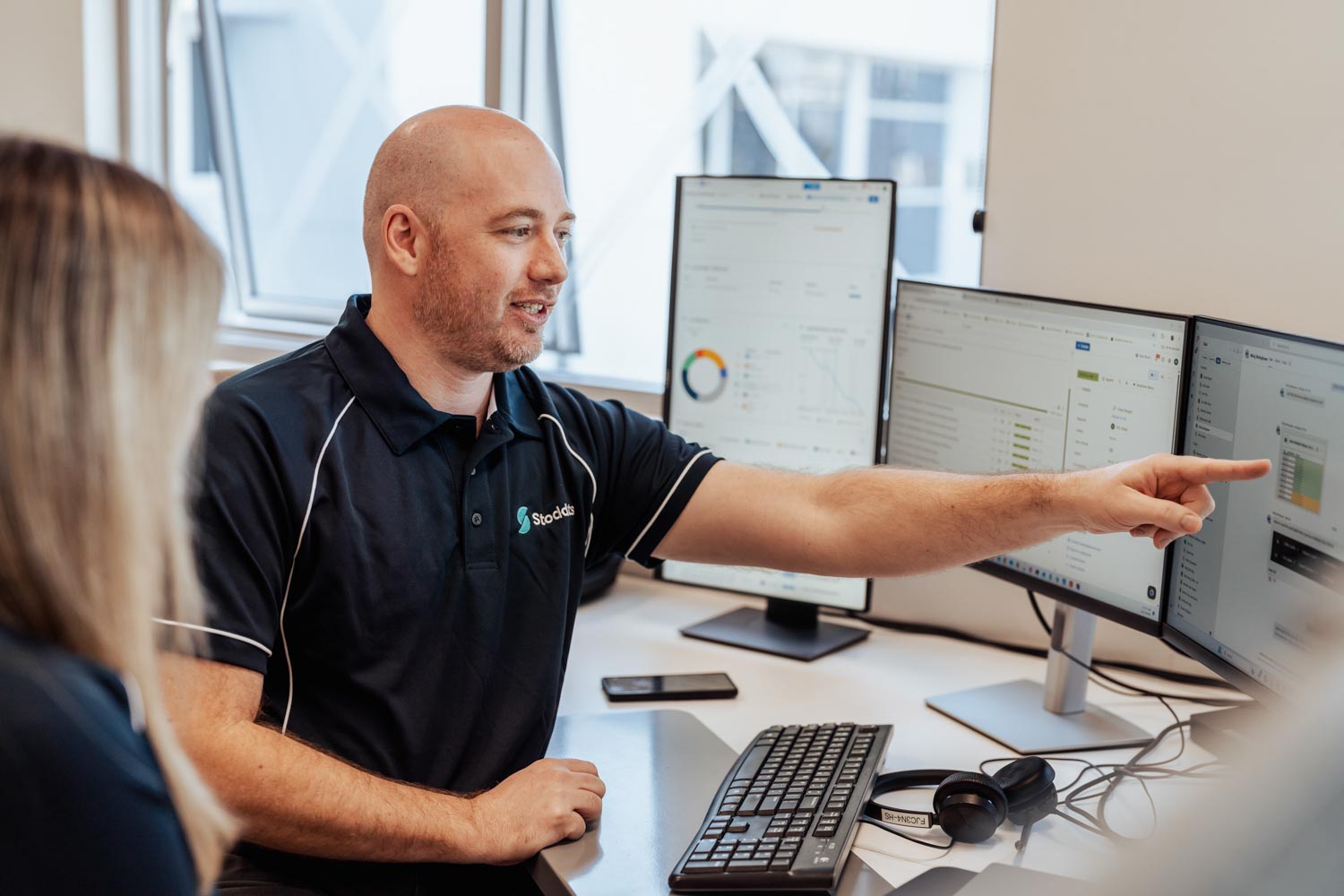 Man in Stoddarts shirt points at computer screens while discussing with a colleague in an office.