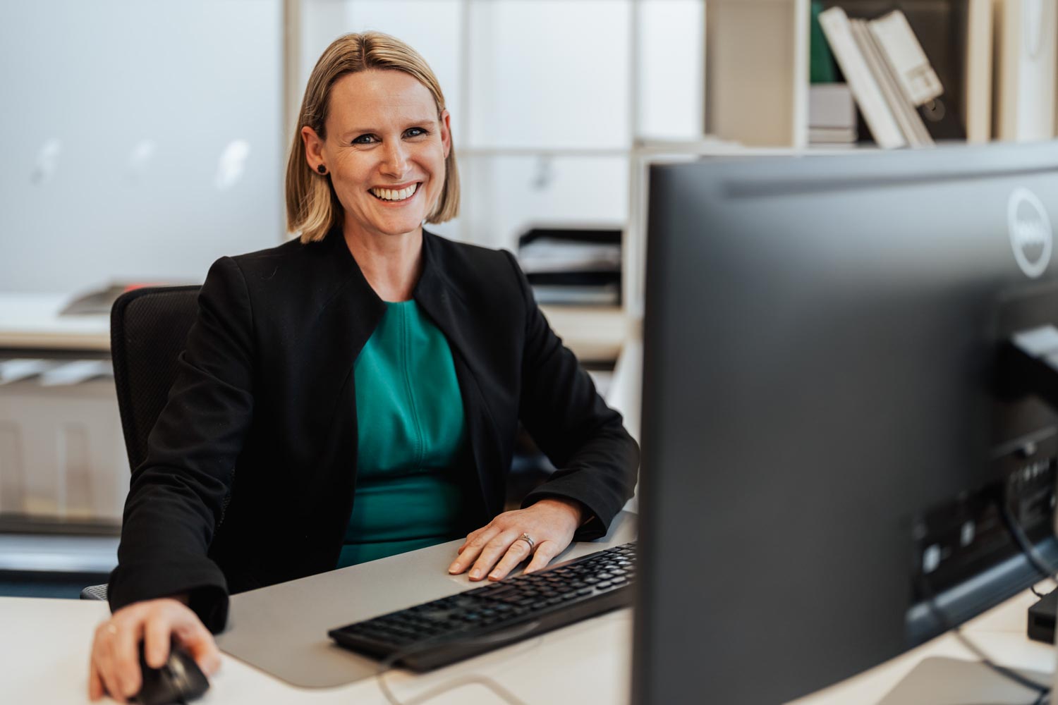 Smiling woman in a black jacket and teal top sitting at a desk using a computer mouse and keyboard.