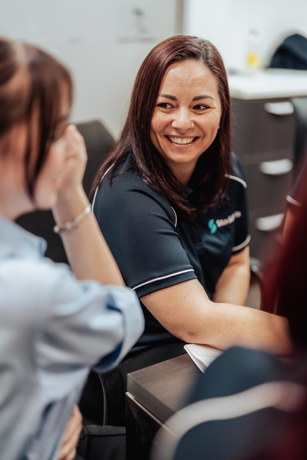 Smiling woman in dark shirt engaged in conversation with another person in an indoor office setting.