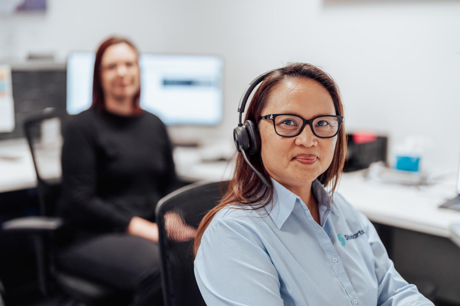 Woman with glasses and headset in blue shirt at office, another person blurred in background.