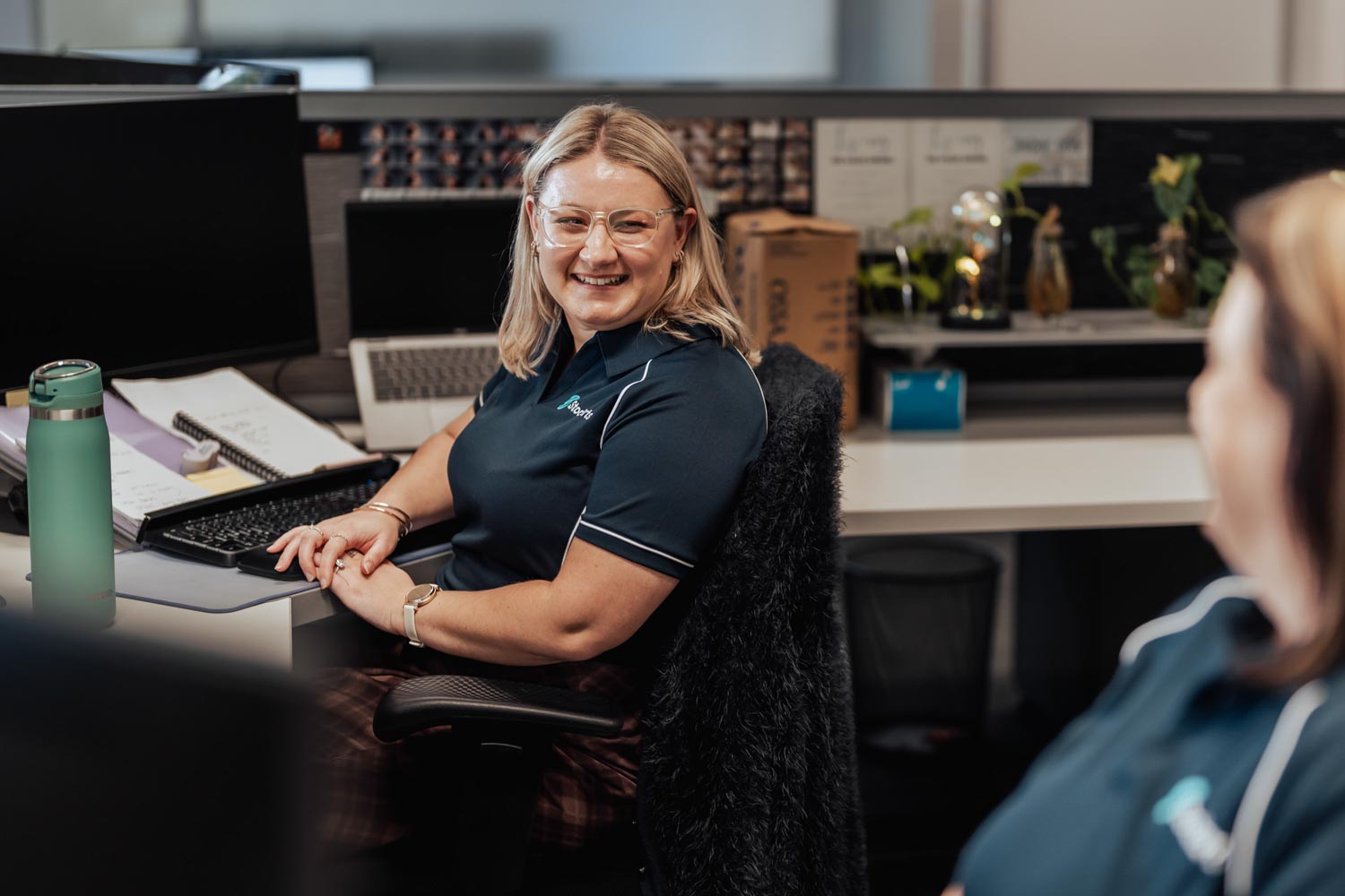 Smiling woman wearing glasses and company polo shirt sitting in an office workspace.