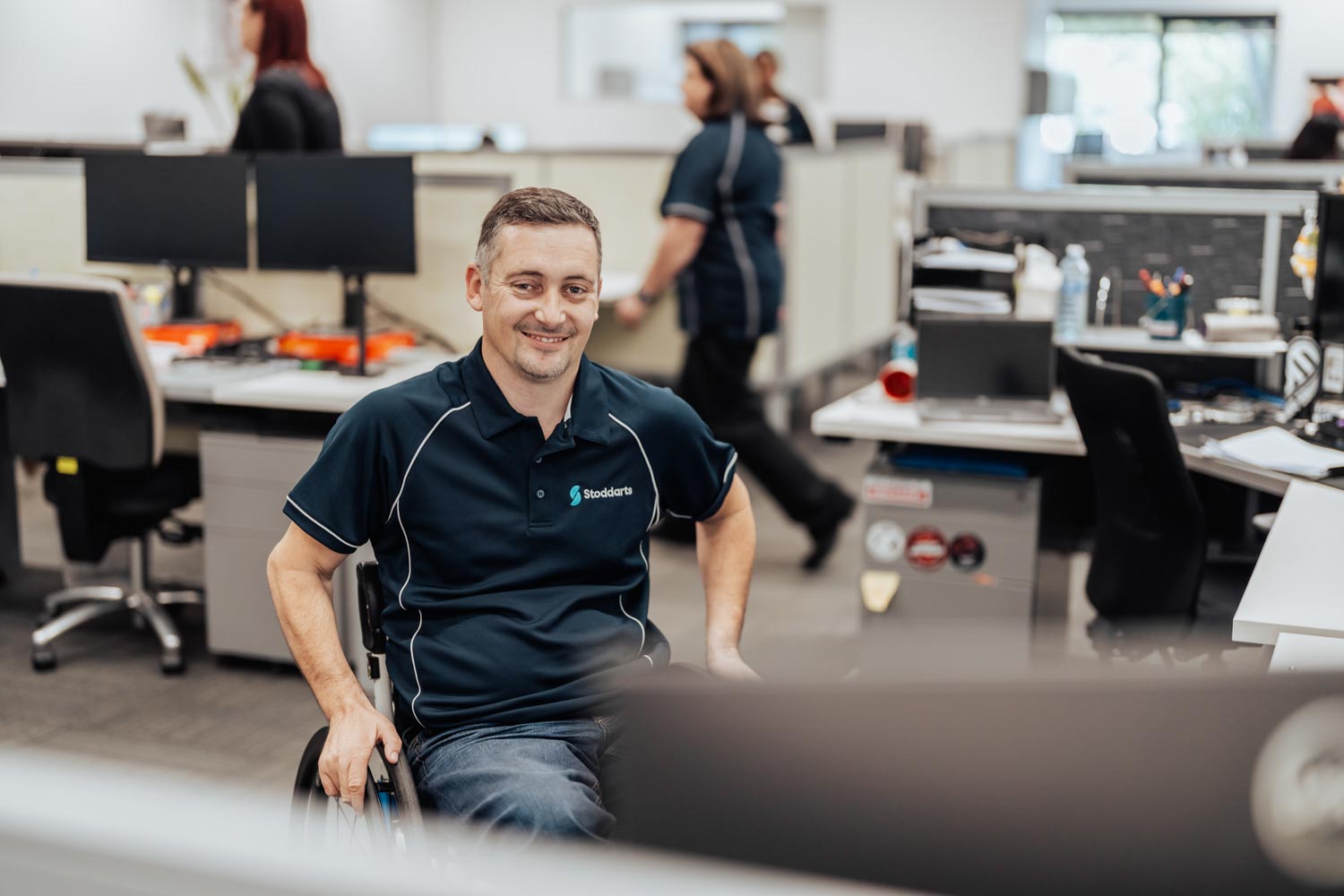 Smiling man in a wheelchair wearing a Stoddarts shirt in a modern office space with coworkers.