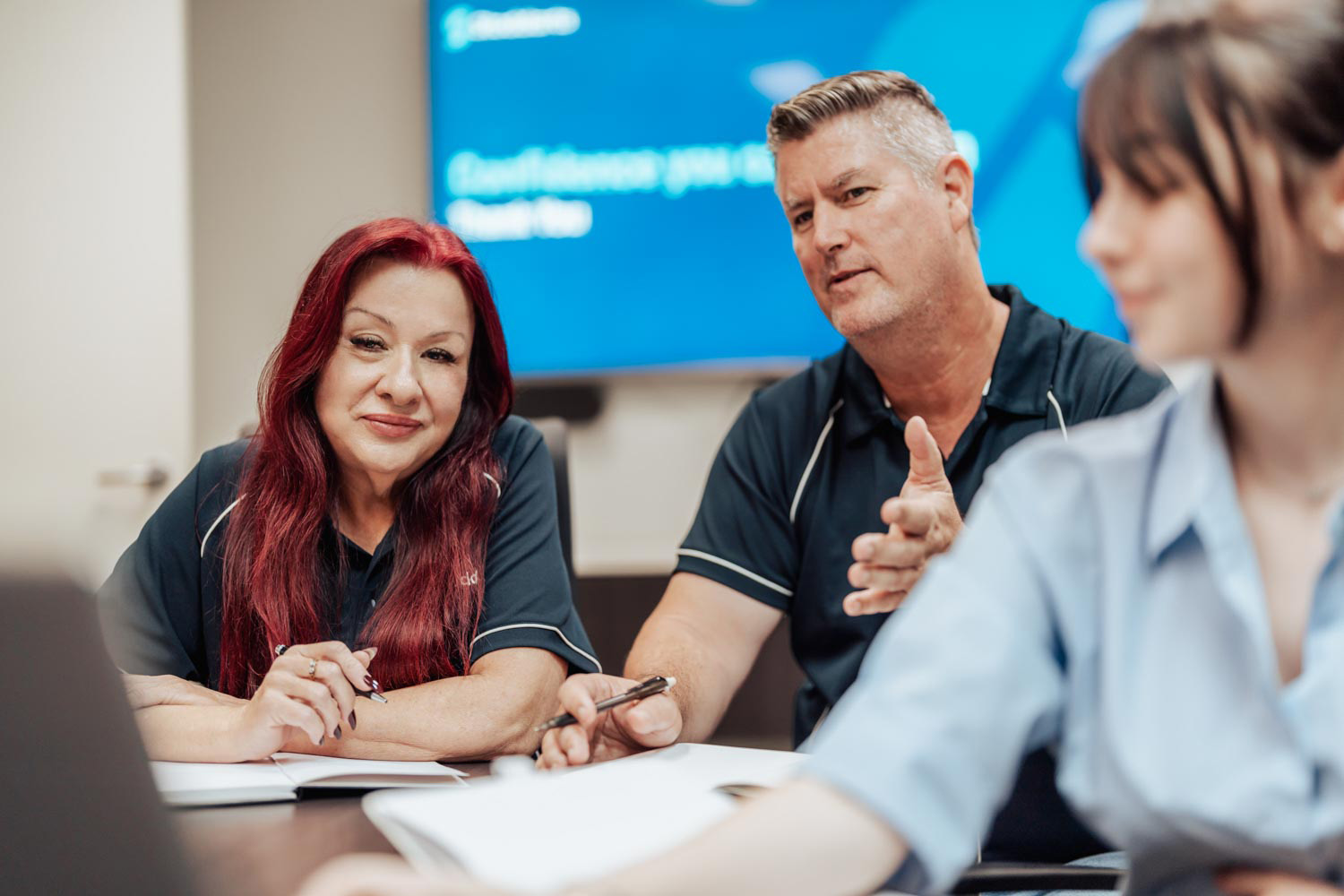 Two adults in black shirts discuss documents with a young woman in a blue shirt in a meeting room.