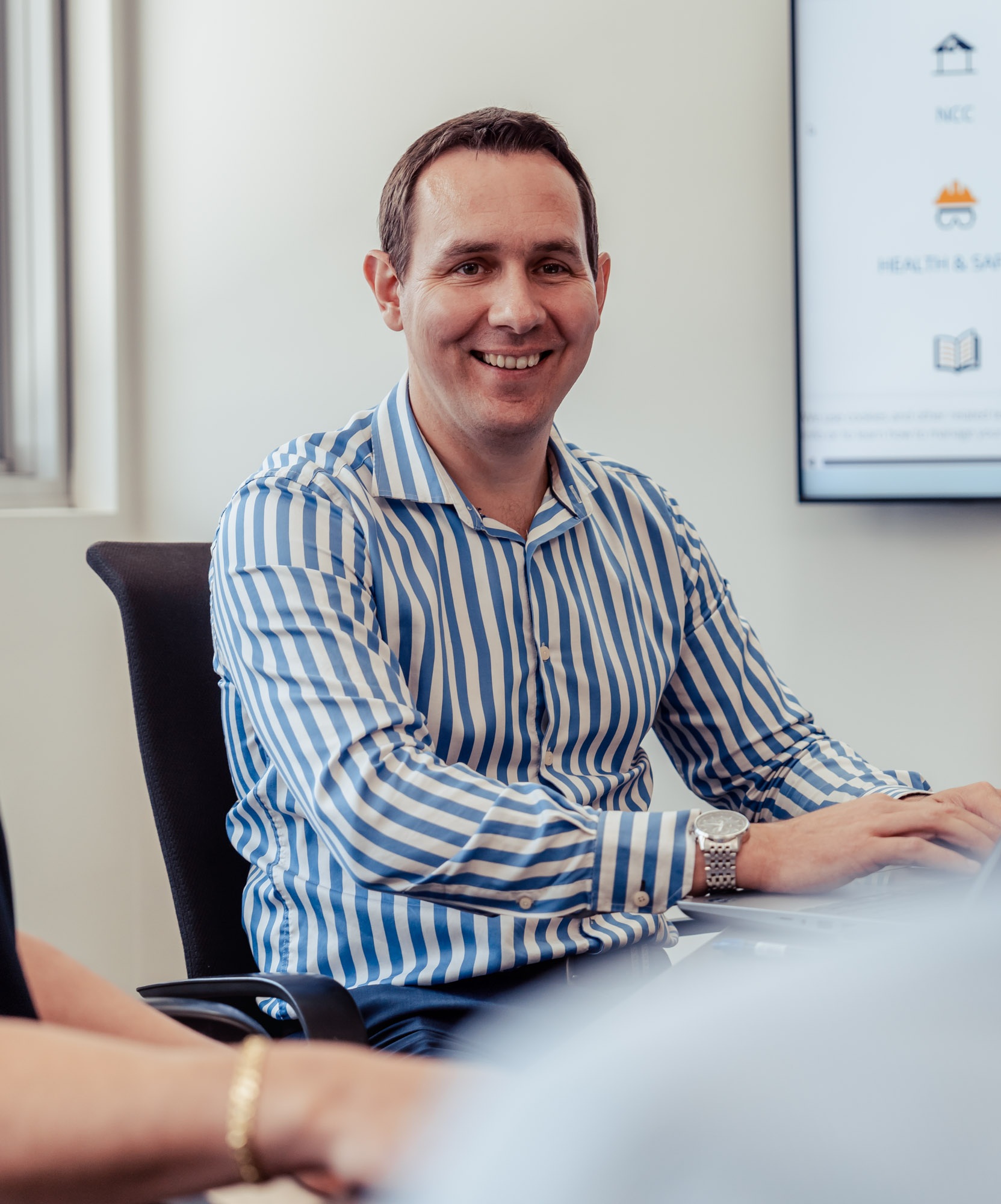 Smiling man in blue and white striped shirt sitting at a desk typing on a laptop.