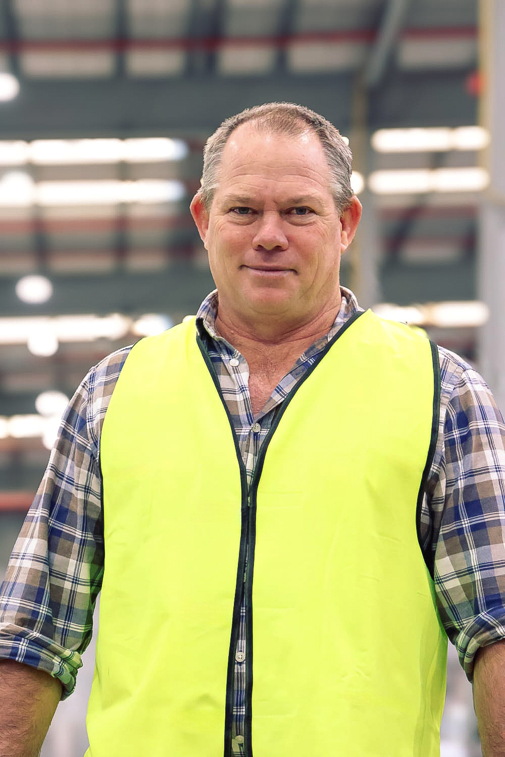 Man in a yellow safety vest and plaid shirt standing in a warehouse or industrial setting.