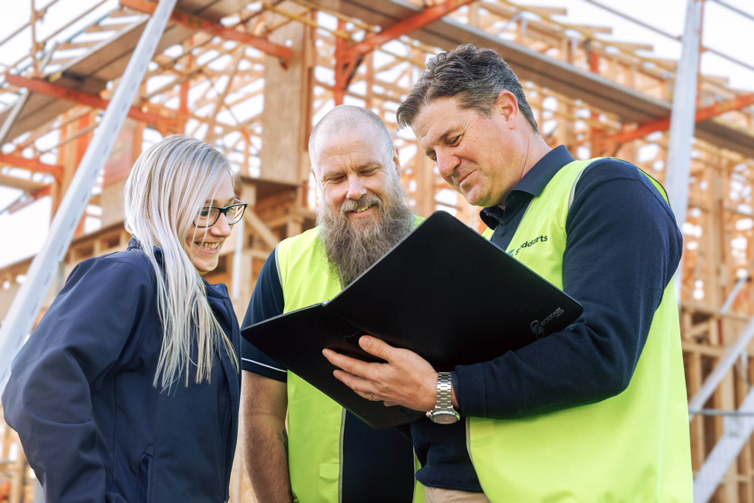 Three construction professionals reviewing documents on site in front of wooden building framing.