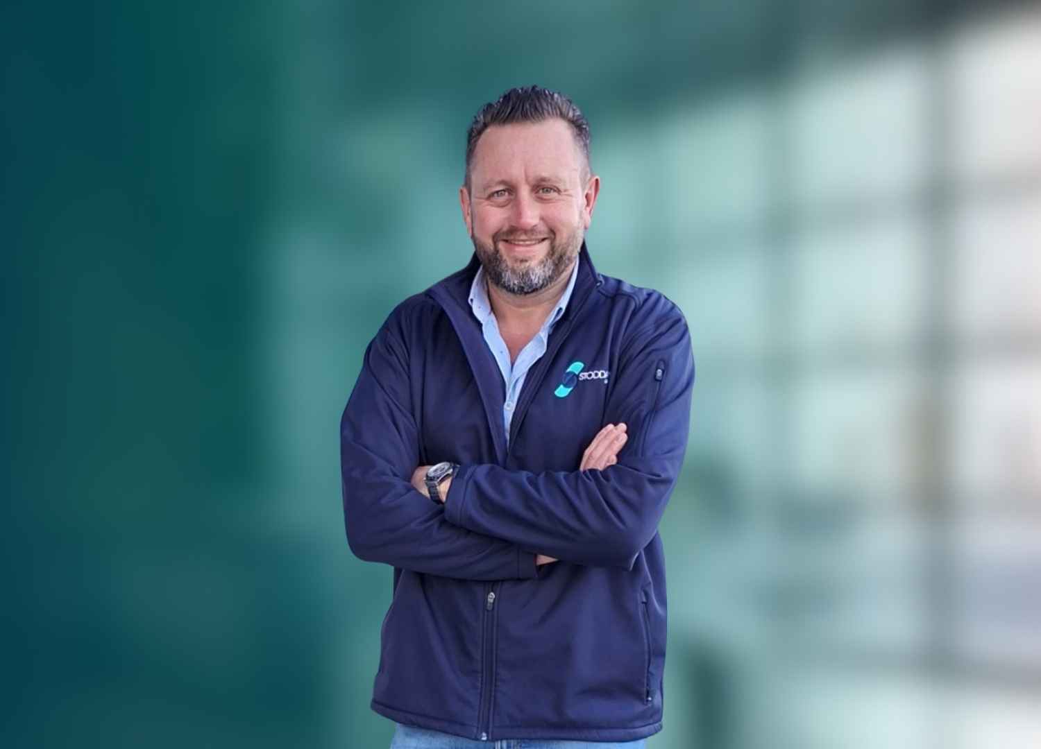 Man with a beard smiling and crossing arms, wearing a navy jacket with a logo over a light blue shirt.