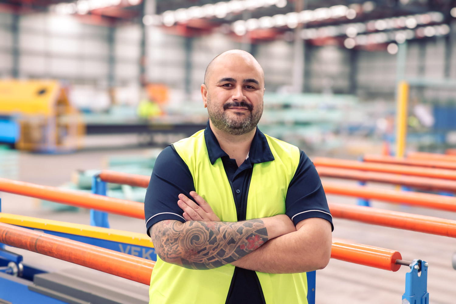 Confident factory worker with tattooed arms and yellow safety vest standing amid orange rails.