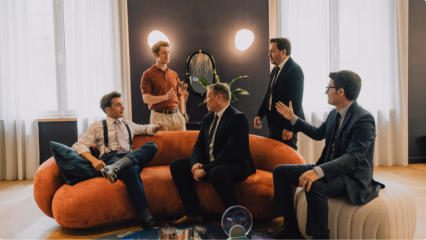Cinq hommes en tenue professionnelle discutent dans un salon moderne, trois assis sur un canapé orange et un pouf, deux debout près d'une plante décorative.
