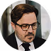 Homme brun avec lunettes et barbe portant un costume et une cravate, regardant vers le bas.