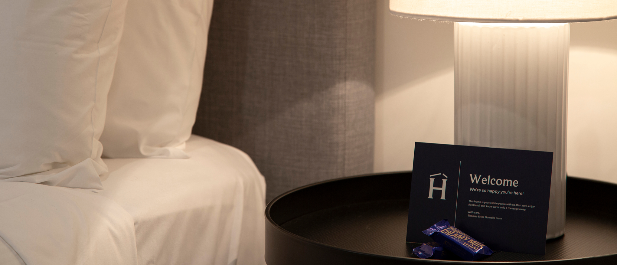 White pillows on a bed next to a bedside table with a lamp, a Homello welcome card, and chocolate bars.