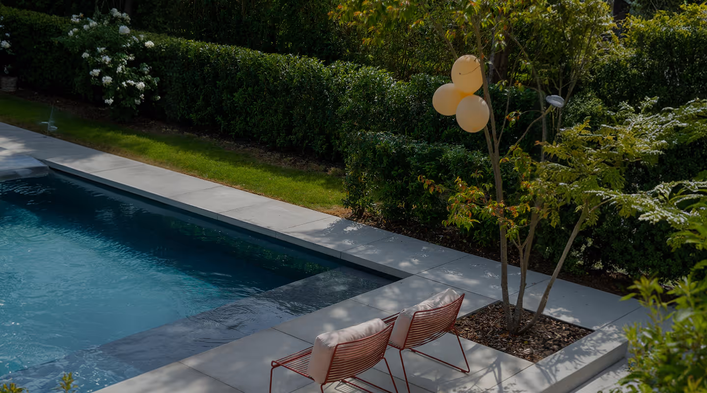 Two red chairs with white cushions by a swimming pool next to a small tree with three yellow balloons.