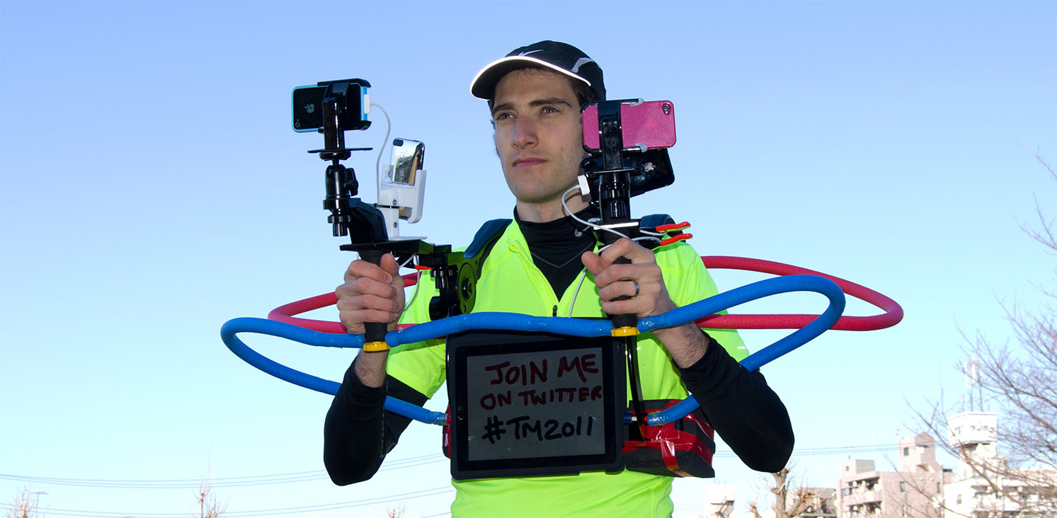 Joseph Tame, in neon green shirt holding dual smartphone mounts with an iPad on his chest displaying 'Join me on Twitter #TM2011'.