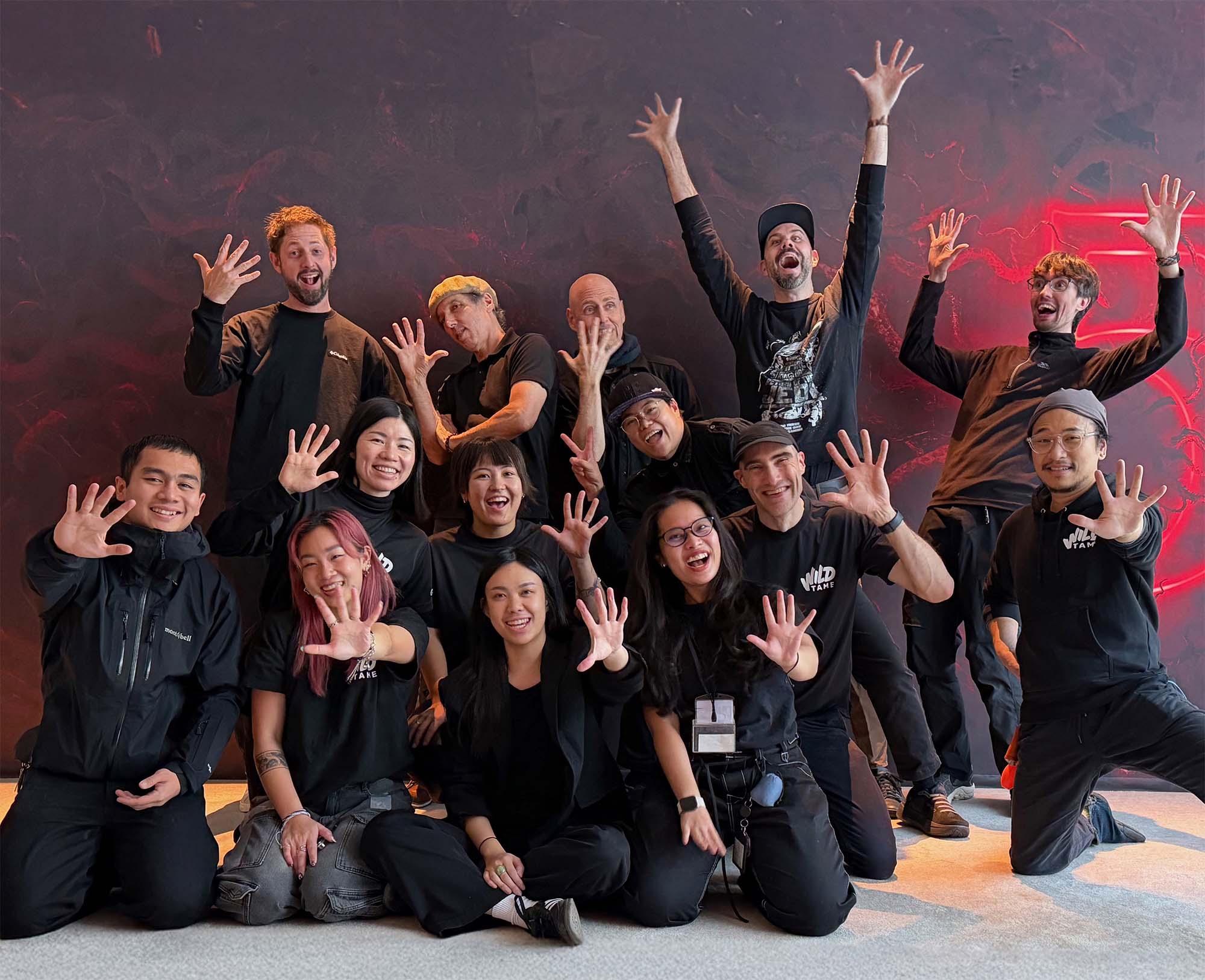 Wild Tame team smiling, posing indoors against a dark red wall, many showing five fingers on one hand.