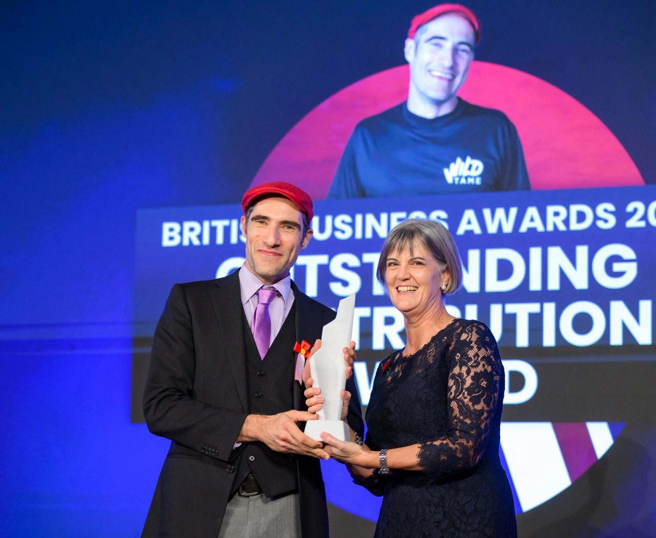 Joseph Tame, accepting an award from the British Ambassador to Japan, holding a white trophy at the British Business Awards ceremony on stage.