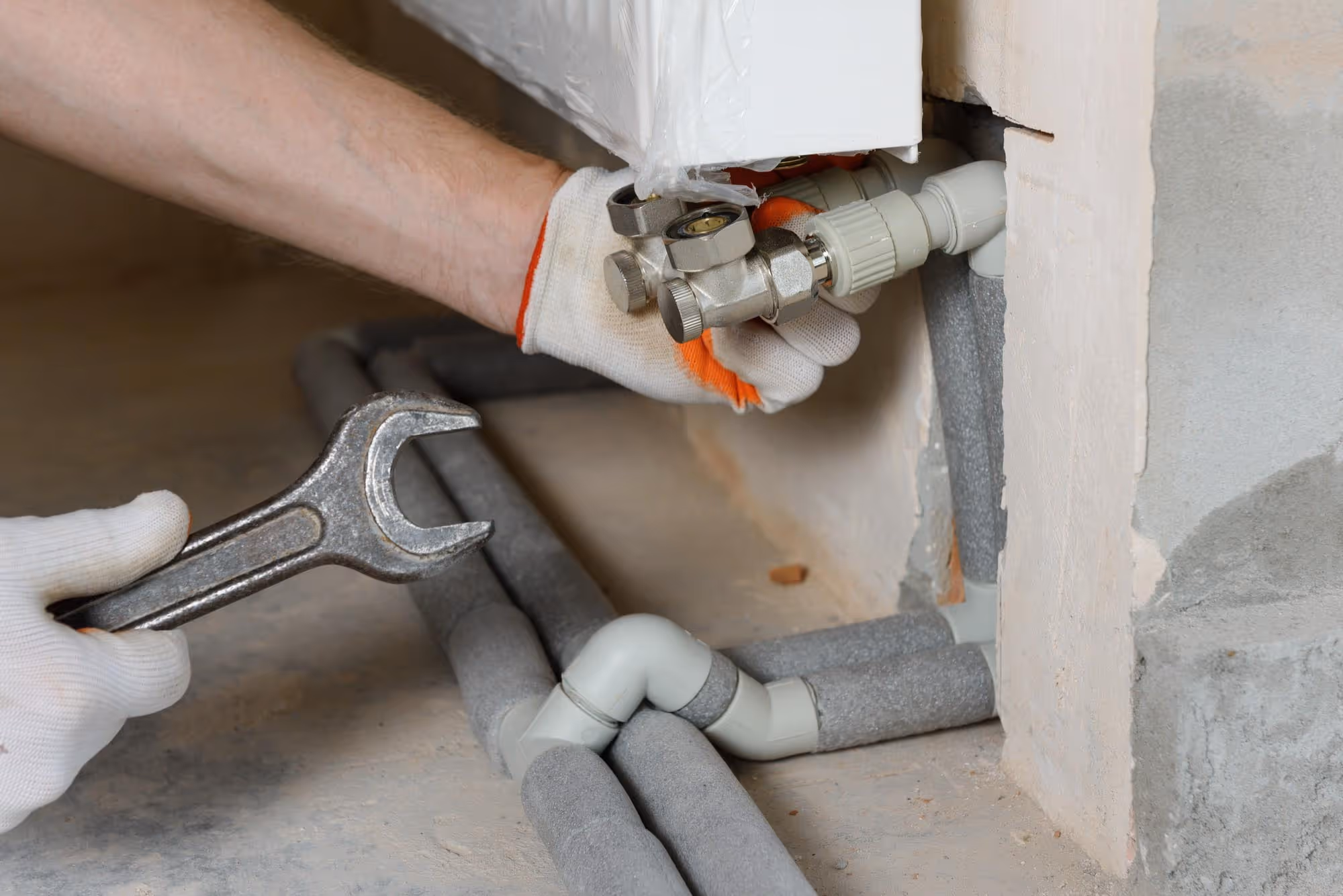 Person tightening a pipe connection near a radiator with a wrench, wearing gloves.