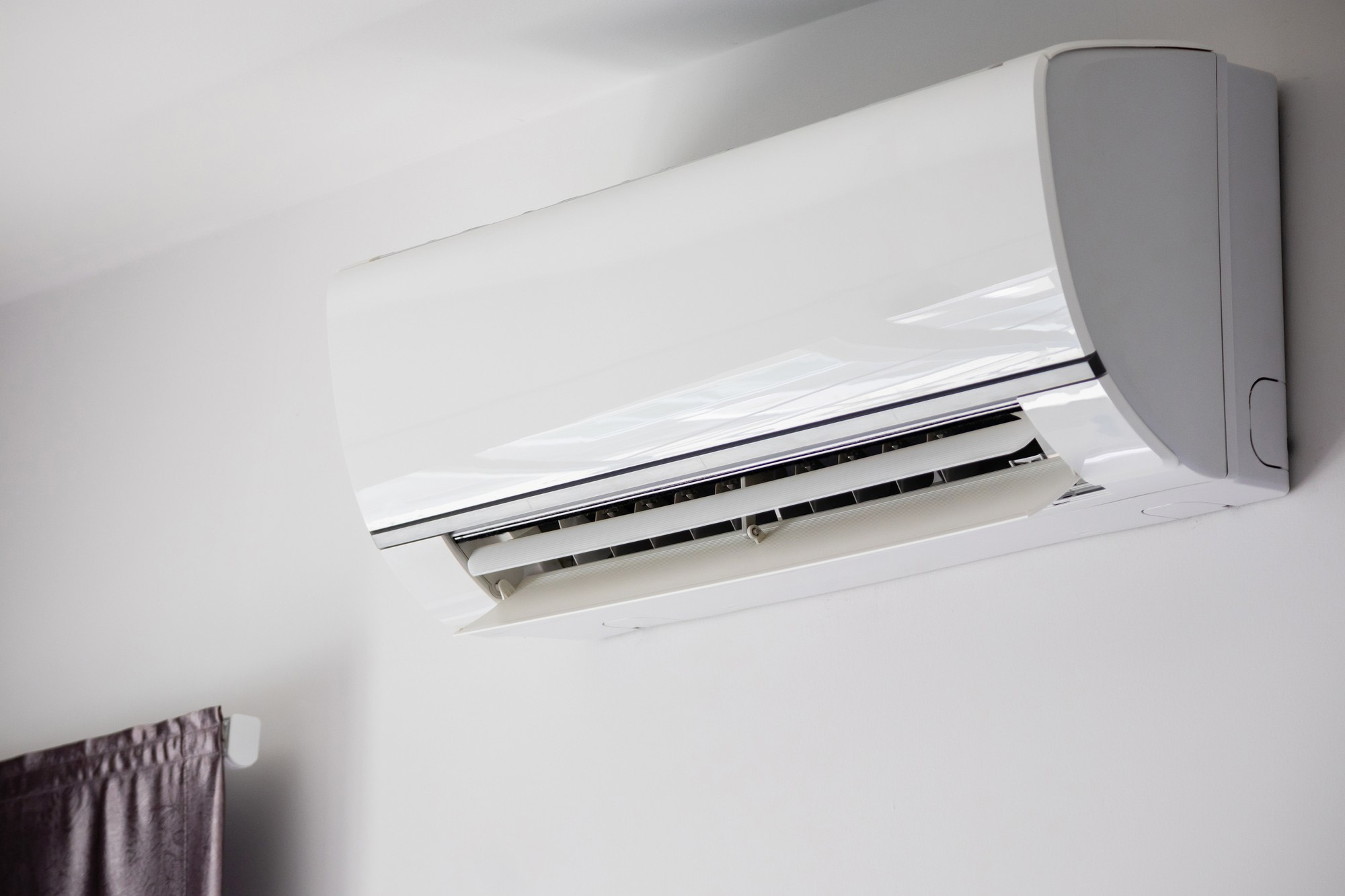 A wall-mounted air conditioning unit with open vents is installed above a curtain.